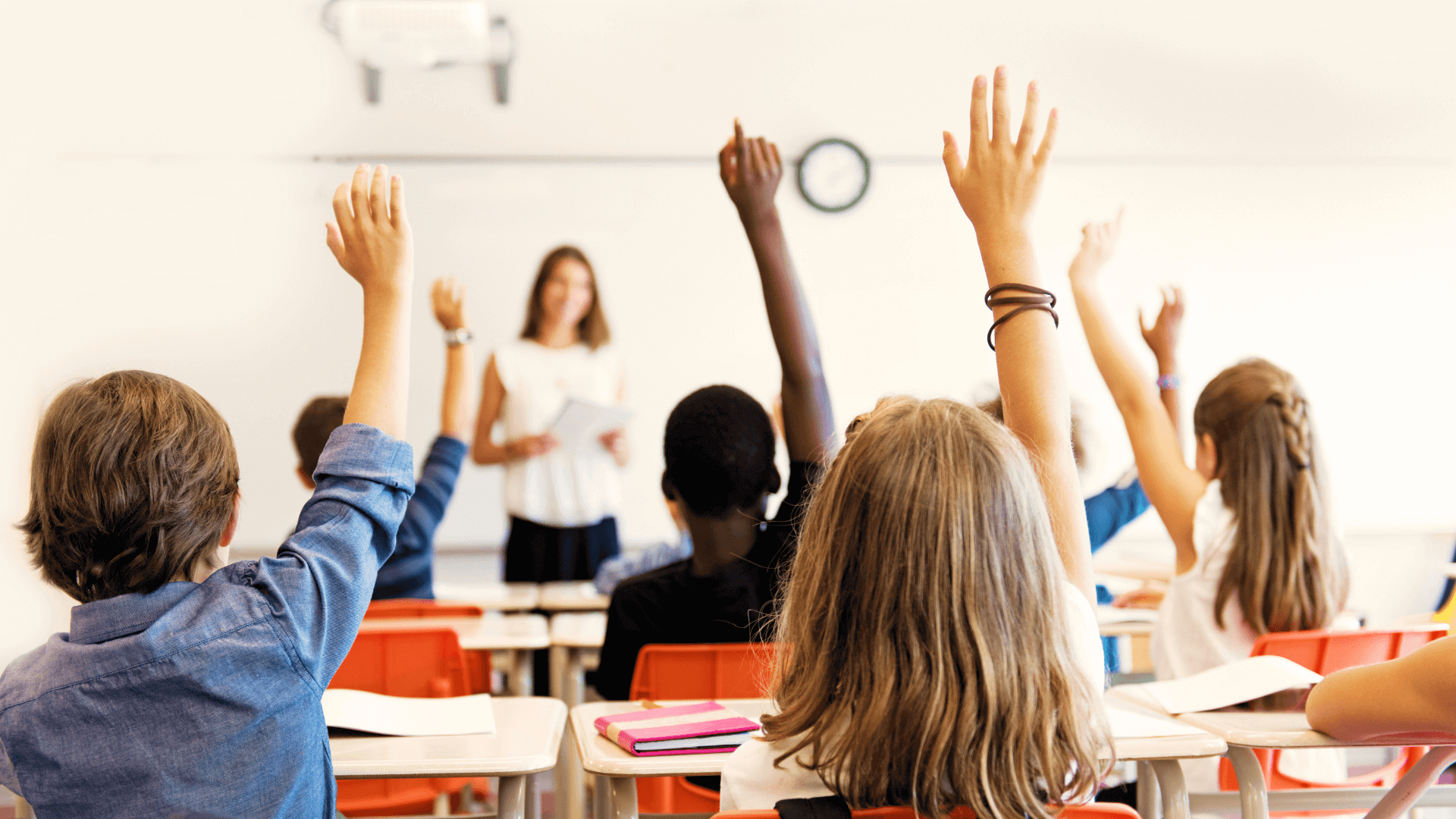 A classroom scene with students seated at desks, eagerly raising their hands to answer a question. A teacher stands at the front near a whiteboard, holding a book. The room has a clock on the wall and bright orange chairs.