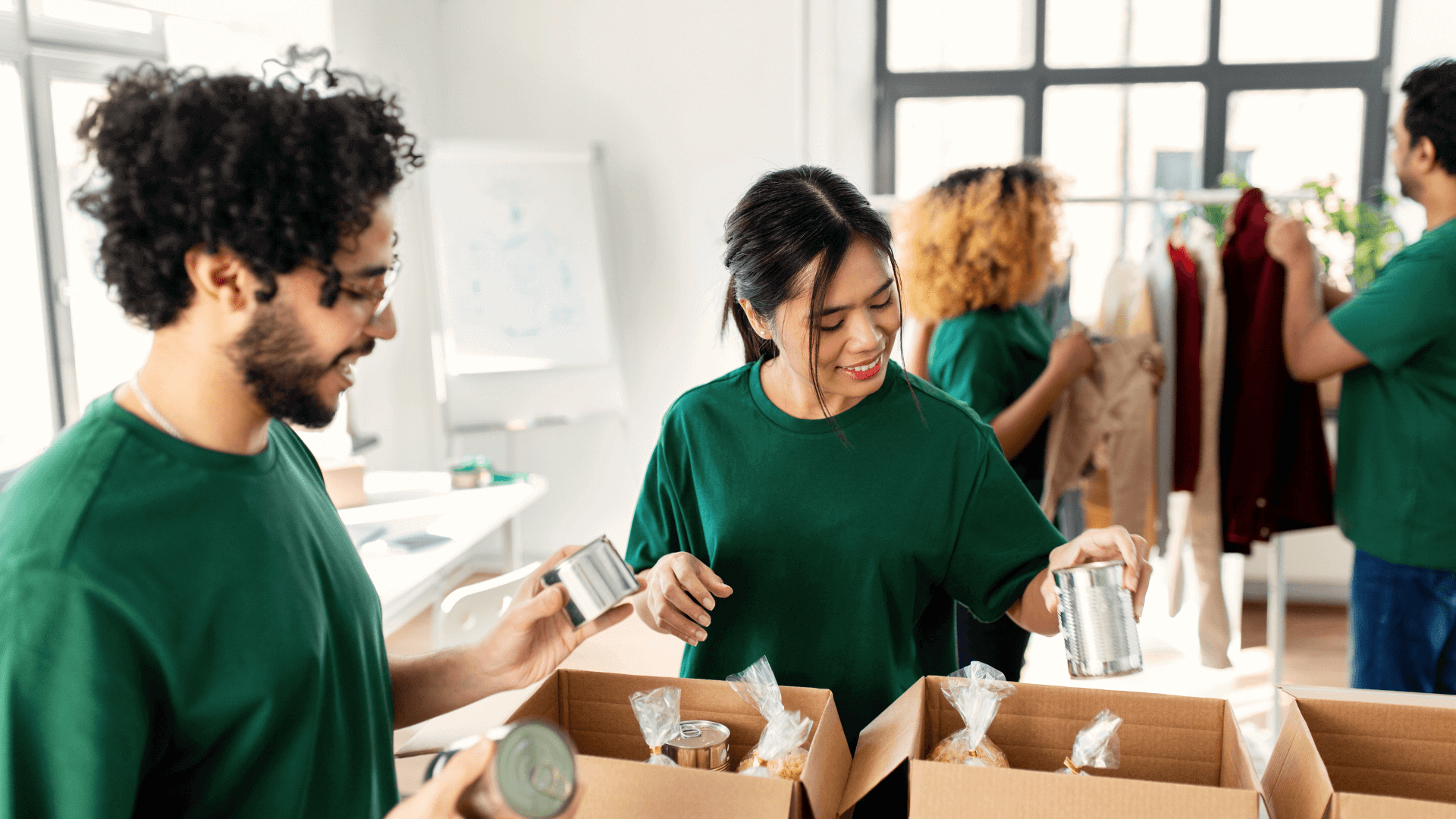 A group of volunteers in green shirts collaboratively packs canned goods and other supplies into cardboard boxes, with clothing items visible in the background, symbolizing a nonprofit organization's efforts to support community welfare and resource distribution.