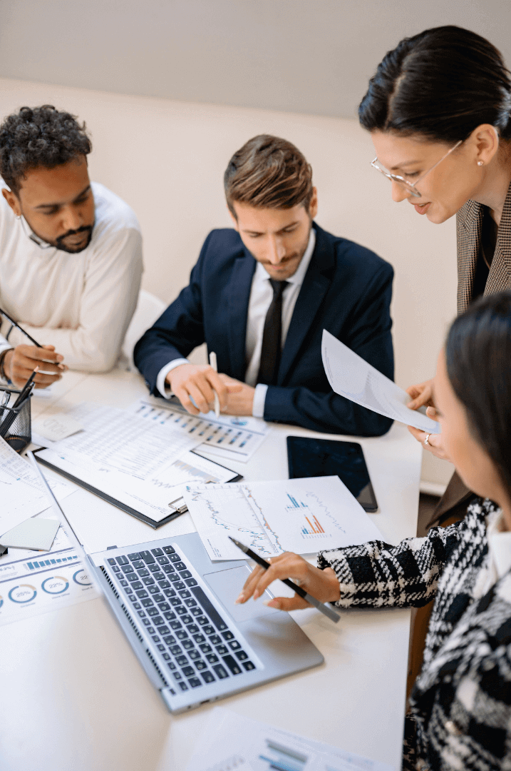 A team of professionals in business attire collaborates around a table, analyzing charts, graphs, and documents on paper and digital devices, symbolizing a strategic approach to grant writing, planning, and sustainable funding solutions. The scene reflects an organization’s journey from launching initiatives to ensuring long-term sustainability through purposeful leadership and structured growth.